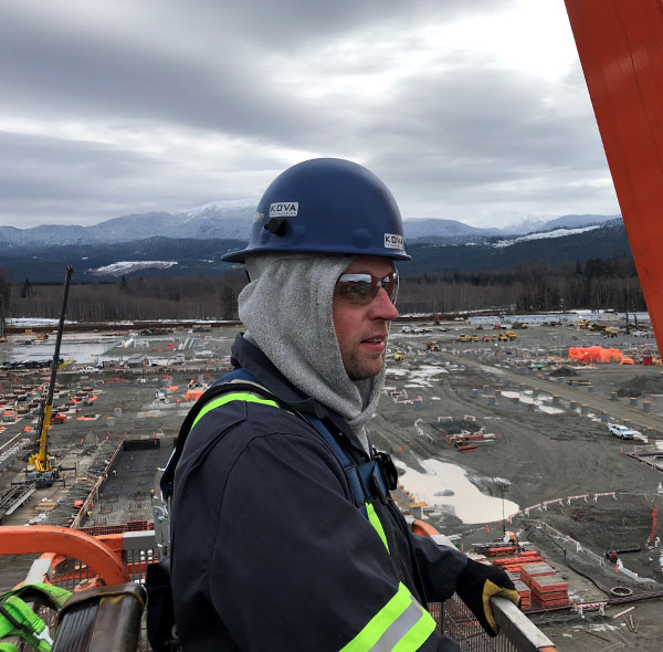 Man with blue hard hat & sunglasses stands on the top of a metal industrial strucutre and looks off into the distance